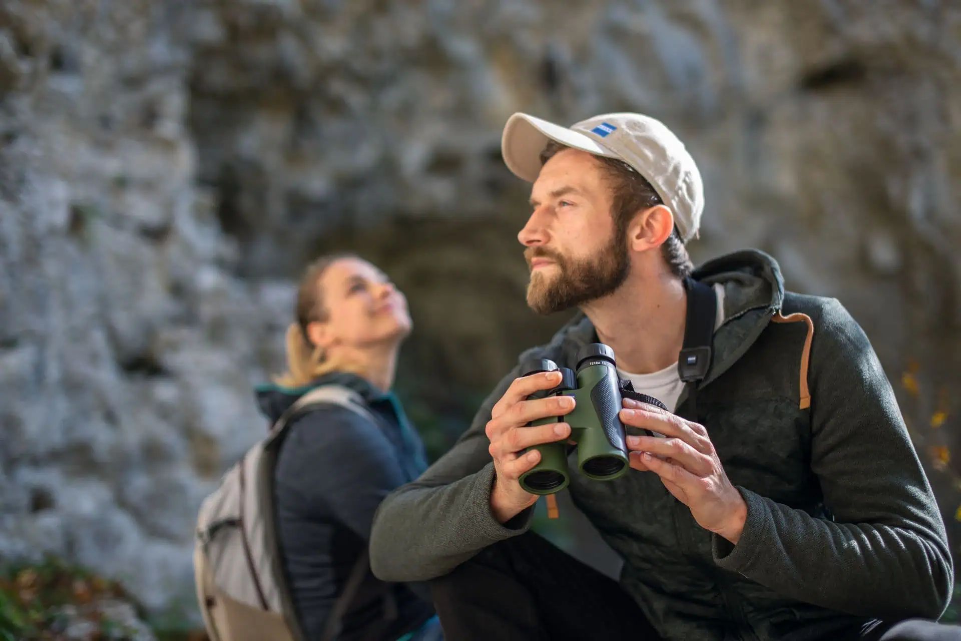 Man in a cap holding green binoculars, a woman by rocks in the background