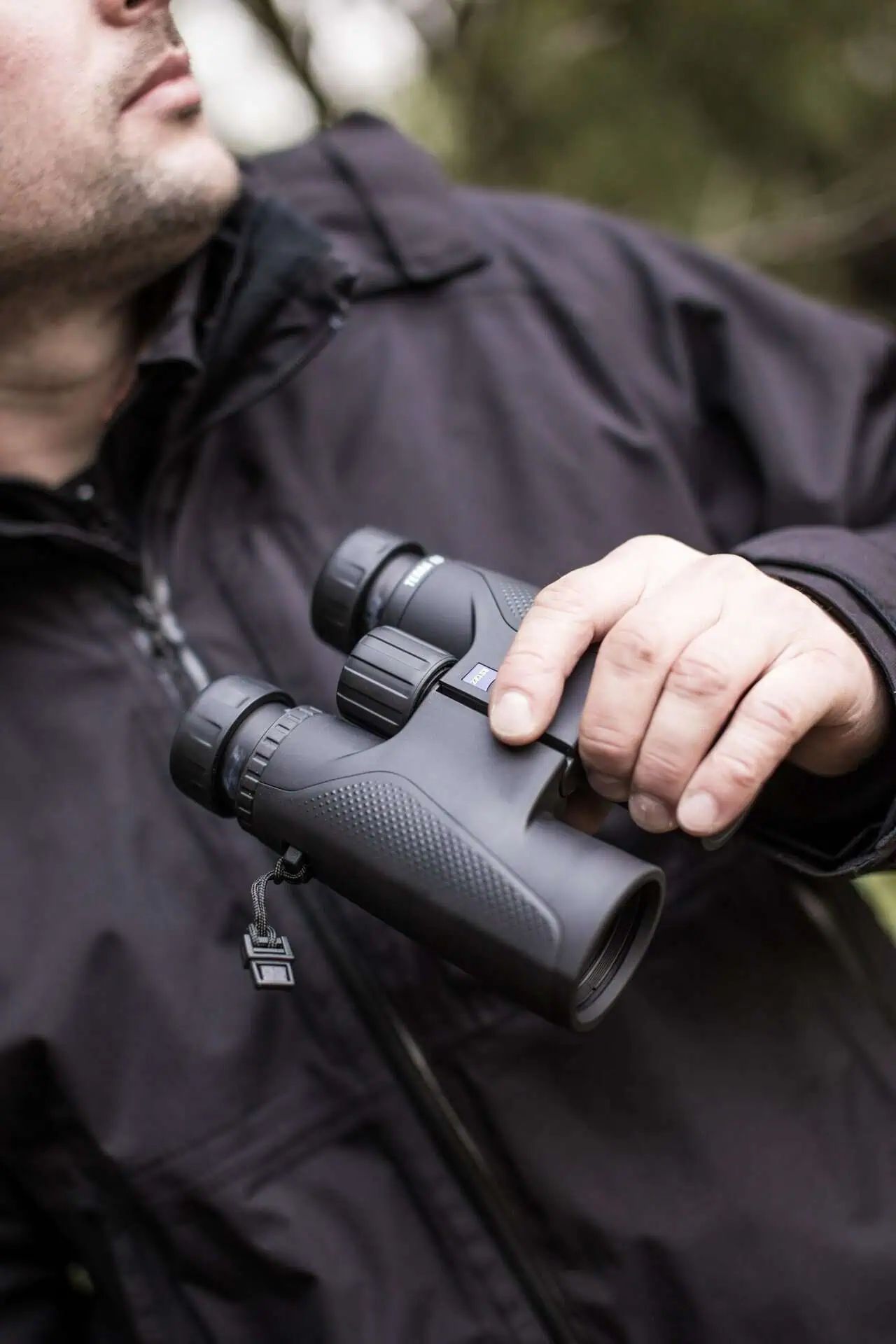 Personne en veste sombre tenant des jumelles noires avec le logo Zeiss