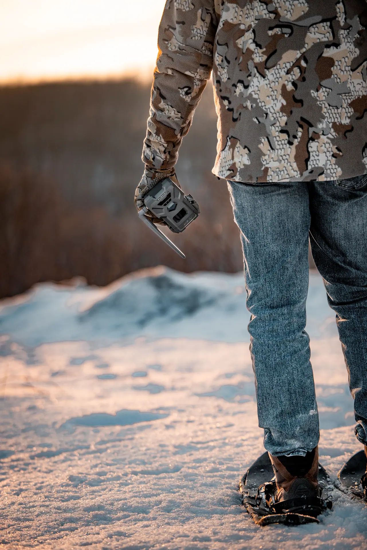 Person in camo jacket and jeans holding a wildlife camera, standing on snowy ground